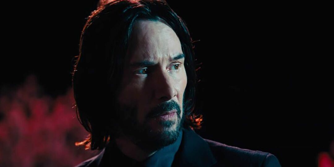 John Wick parecendo arrumado, parado em um quarto escuro.